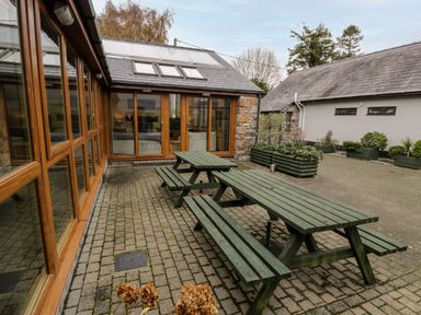An outdoor patio area with green wooden picnic tables and benches next to a building with large wooden framed windows at Ddol Helyg Barn in Llanrug