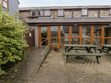 A paved outdoor patio area with green picnic benches and a potted plant in front of a stone building with wooden framed windows at Ddol Helyg Barn in Llanrug