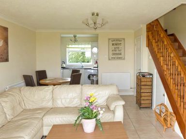 A living room with a beige sectional sofa a wooden coffee table with a flower pot a dining table with four chairs a staircase and a kitchen in the background at Seascope in Kessingland near Lowestoft