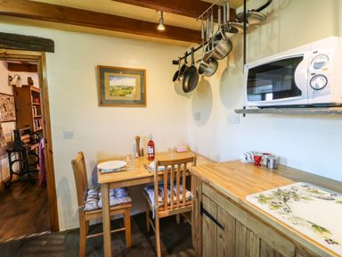A small kitchen area with a wooden table and chairs set for two a microwave on a wooden counter and pots hanging from the ceiling at Cherry Tree Cottage in Soulby near Kirkby Stephen