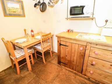 A small kitchen corner with a wooden table and two chairs set with plates glasses and a bottle of wine a wooden cabinet with drawers and a microwave on a shelf above at Cherry Tree Cottage Soulby near Kirkby Stephen
