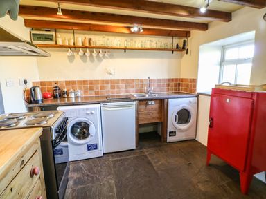 A kitchen with a stove washing machines a refrigerator and a shelf with bottles and mugs at Cherry Tree Cottage Soulby near Kirkby Stephen