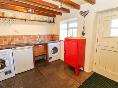 A kitchen with a washing machine dishwasher red cabinet and sink under a shelf with glass bottles at Cherry Tree Cottage Soulby near Kirkby Stephen