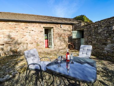 An outdoor patio with a stone wall a glass table with two cushioned chairs a bottle of wine and two glasses at Cherry Tree Cottage Soulby near Kirkby Stephen