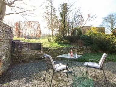 An outdoor patio with two cushioned chairs and a table with a bottle and glasses surrounded by stone walls at Cherry Tree Cottage in Soulby near Kirkby Stephen