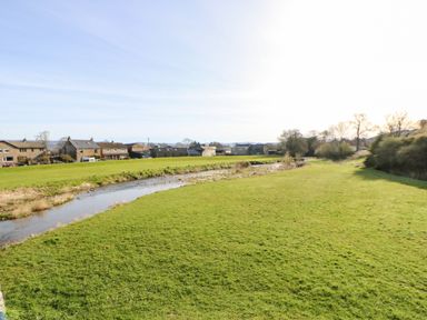 A grassy field with a small river running through it and houses in the background at Cherry Tree Cottage in Soulby near Kirkby Stephen