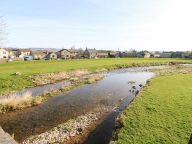 A river flowing through a grassy field with houses in the background at Cherry Tree Cottage in Soulby near Kirkby Stephen