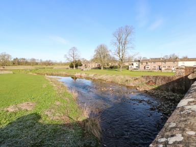 A river flowing through grassy fields with trees and stone houses in the background at Cherry Tree Cottage in Soulby near Kirkby Stephen
