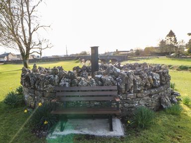 An outdoor bench in front of a circular stone structure on a grassy area with trees and houses in the background