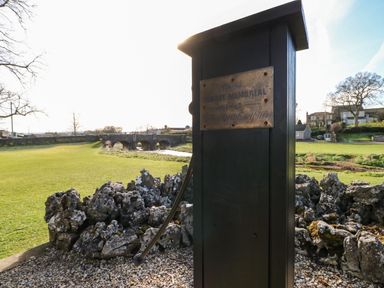 A metal post with a plaque in a grassy area near a stone wall and a stone bridge in the background
