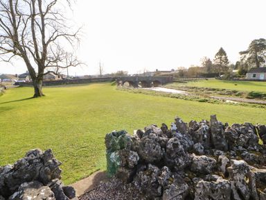 An outdoor scene with a stone wall in the foreground a large tree a grassy area a stone bridge over a stream and houses in the background at Cherry Tree Cottage in Soulby near Kirkby Stephen
