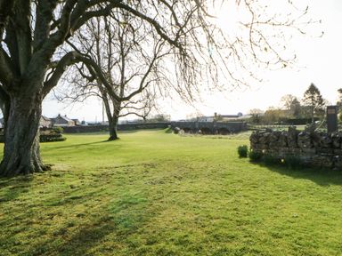 A grassy open area with trees a stone wall and a bridge with houses in the background at Cherry Tree Cottage in Soulby near Kirkby Stephen