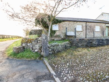 A stone cottage with three windows and a bench outside near a cobblestone path and a stone wall with a tree at Cherry Tree Cottage in Soulby near Kirkby Stephen