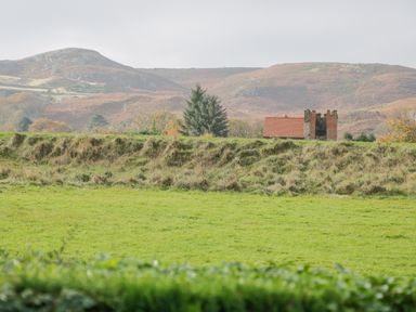 A green field with a hedge and a small house with a red roof and chimney stacks in front of hills at Mountain View in Conwy