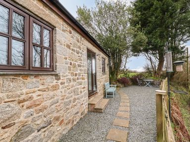 An outdoor seating area with gravel ground a stone building with windows and door benches and a table with chairs at Brocks Barn Sweetshouse near Lostwithiel