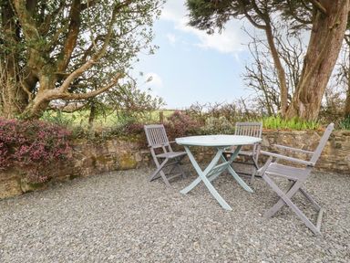An outdoor seating area with a round table and three chairs on gravel next to a stone wall and trees at Brocks Barn Sweetshouse near Lostwithiel