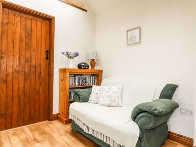 A small living room with a green sofa covered by a white throw and patterned cushions next to a wooden shelf with books and a vase of flowers at Brocks Barn Sweetshouse near Lostwithiel