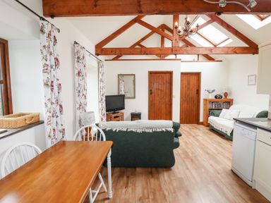 A living room with wooden beams two green sofas a wooden dining table and floral curtains at Brocks Barn Sweetshouse near Lostwithiel