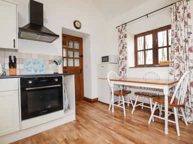 A kitchen with a wooden door oven and dining table with chairs near a window with floral curtains at Brocks Barn Sweetshouse near Lostwithiel