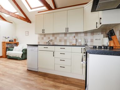 A kitchen with white cabinets black countertop and wooden beams on the ceiling with a green armchair and wooden cabinet in the background at Brocks Barn in Sweetshouse near Lostwithiel
