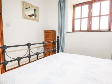 A bedroom with a metal bed frame wooden chest of drawers a window with wooden framing and curtains and a wall mirror at Brocks Barn in Sweetshouse near Lostwithiel