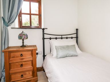 A bedroom with a single bed white bedding a wooden nightstand with a stained glass lamp and a window with blue curtains at Brocks Barn Sweetshouse near Lostwithiel