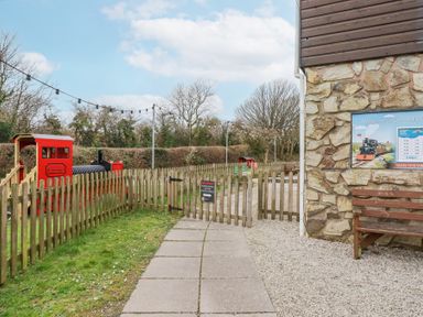 A small outdoor train track with a red train and a wooden fence near a stone building with a bench and a train timetable at The Station Flat in St Newlyn East near Newquay