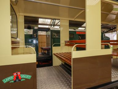The interior of a train carriage with wooden benches and a view of a green locomotive outside at Lappa Valley