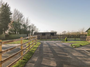 A fenced outdoor area with a paved path leading to three small wooden shelters surrounded by grass and trees at The Log Cabin in Bushton near Royal Wootton Bassett
