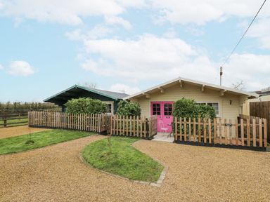 A gravel driveway leading to two small cabins with wooden fences and shrubs in front at The Log Cabin in Bushton near Royal Wootton Bassett