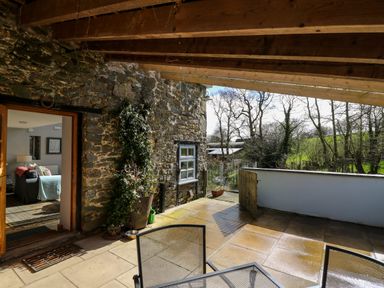 A stone patio with metal chairs and a wooden pergola next to a stone wall and an open door showing a living room at The Barn at Glanoer Hundred House near Builth Wells