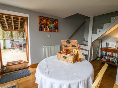 A dining area with a round table covered with a white cloth holding a basket of food and a bottle near a staircase and an open door showing an outdoor seating area at The Barn at Glanoer in Hundred House near Builth Wells