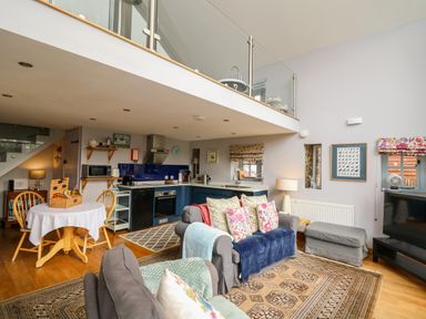 An open living area with a kitchen, dining table with four chairs, sofas with cushions, a TV, and a mezzanine at The Barn at Glanoer Hundred House near Builth Wells