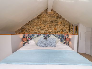 A bed with patterned headboard blue and beige pillows and a light blue blanket under a sloped ceiling and stone wall at The Barn at Glanoer Hundred House near Builth Wells