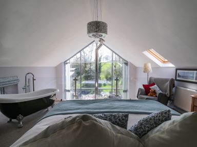 A bedroom with a bed a bathtub an armchair with cushions and a teddy by a large window at The Barn at Glanoer in Hundred House near Builth Wells