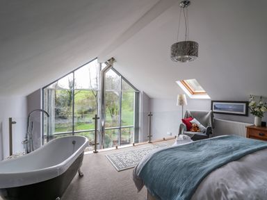 A bedroom with a bathtub near large windows a chair with cushions a bed with a blue blanket and a skylight at The Barn at Glanoer in Hundred House near Builth Wells