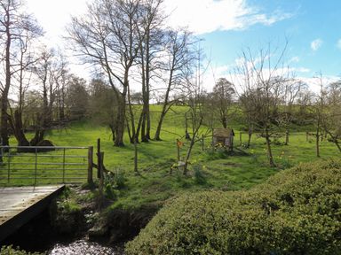 A grassy field with trees a small wooden structure a metal gate and a wooden bridge at The Barn at Glanoer in Hundred House near Builth Wells