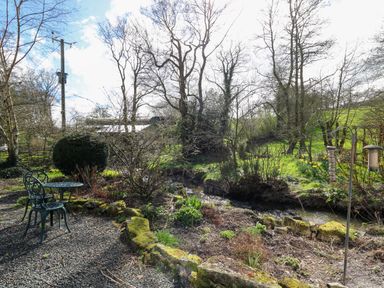 A garden area with a small metal table and two chairs next to a stream and trees at The Barn at Glanoer Hundred House near Builth Wells