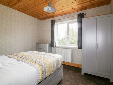 A bedroom with a bed a gray wardrobe a dresser window with curtains and a wooden ceiling at Lodge One in Brighton near Summercourt