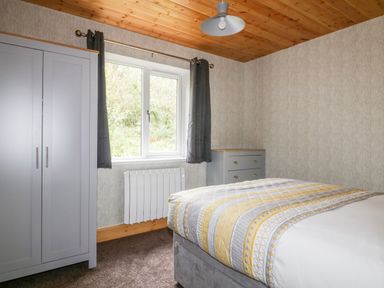 A bedroom with a bed covered with patterned bedding a grey wardrobe a chest of drawers and a window with dark curtains at Lodge One Brighton near Summercourt