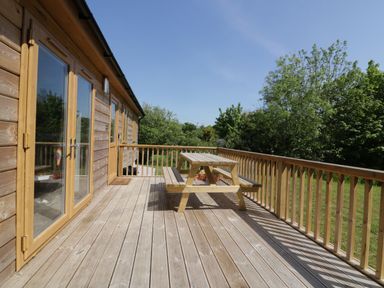 A wooden deck with a picnic table next to a wooden building with glass doors and windows surrounded by trees at Lodge One Brighton near Summercourt