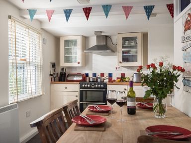 A dining table set with red plates and wine glasses in a kitchen with a stove and cabinets at Thimble Cottage in Mevagissey