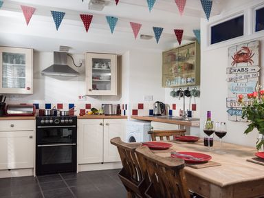 A kitchen with wooden table and chairs red plates and wine glasses on the table kitchen appliances and cabinets with colorful bunting hanging from the ceiling at Thimble Cottage in Mevagissey