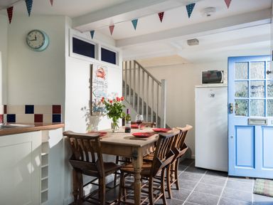 A dining area with a wooden table and chairs a bouquet of flowers and wine on the table a blue door and white refrigerator in the background at Thimble Cottage in Mevagissey