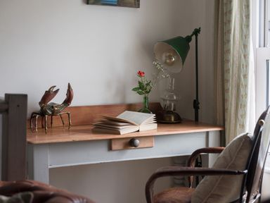 A wooden desk with a metal crab sculpture an open book a green vase with flowers a lamp and a chair at Thimble Cottage in Mevagissey