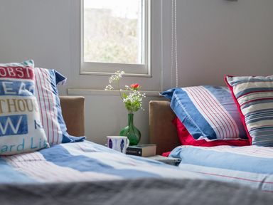 A bedroom with two beds blue striped bedding pillows a green vase with flowers a book and a mug on a small table by a window at Thimble Cottage in Mevagissey