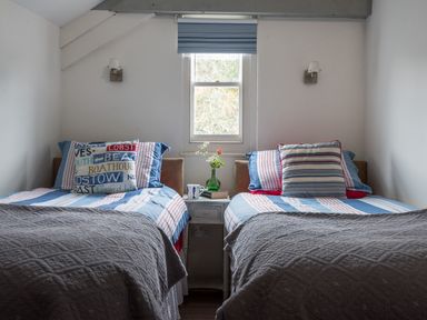 A bedroom with two single beds with striped bedding a window between the beds and a small table with a vase and a mug at Thimble Cottage in Mevagissey