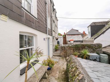 An outdoor stone-paved path with potted plants and a stone bench beside white and brown siding buildings at Thimble Cottage in Mevagissey