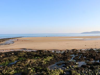 A sandy beach with rocky areas and a person walking dogs near the shore at Beach View in Benllech near Red Wharf Bay