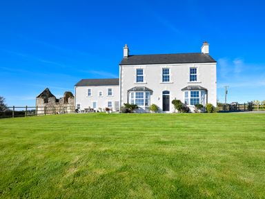 A white two story house with bay windows and a green lawn under a blue sky at Ty Carmel in Rhosneigr